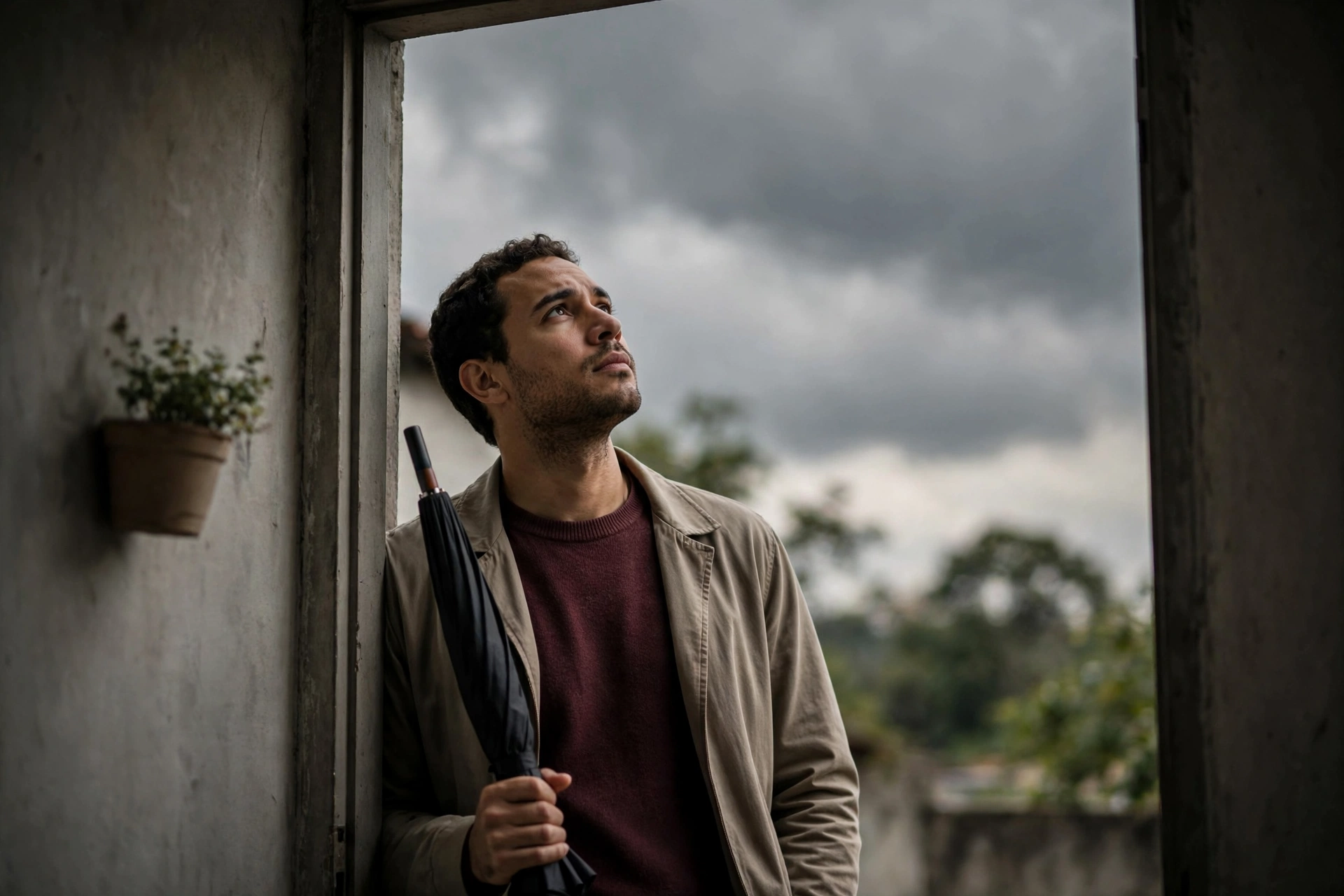 Homem observa o céu nublado na entrada de casa com um guarda-chuva na mão.